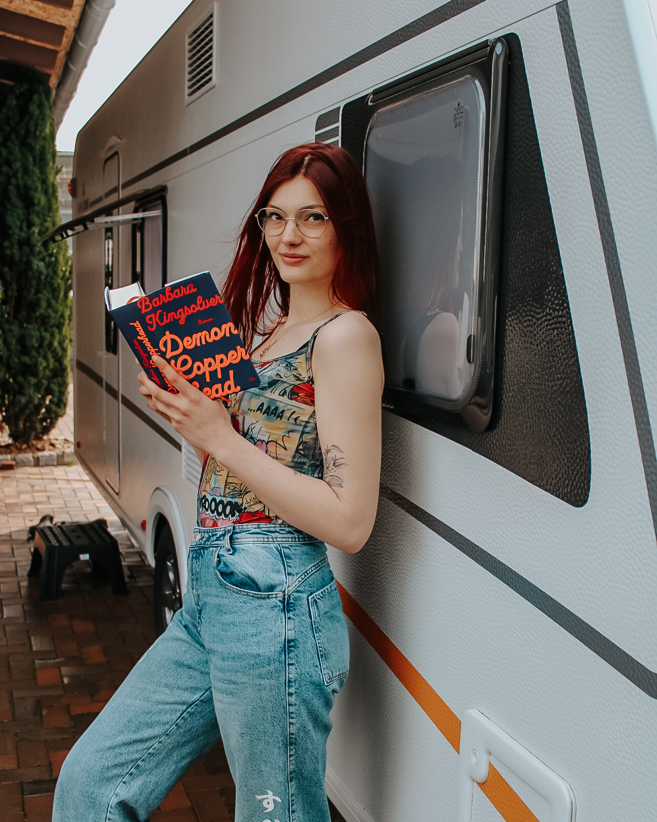 Demon Copperhead Rezension. rothaariges Mädchen hält Buch in der Hand und lehnt an Wohnwagen