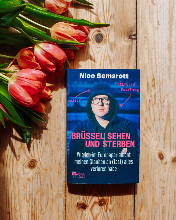Brüssel sehen und sterben von Nico Semsrott. Das Buch liegt mittig auf dem Bild auf einem Holzboden. In der linken oberen Ecke liegen Tulpen